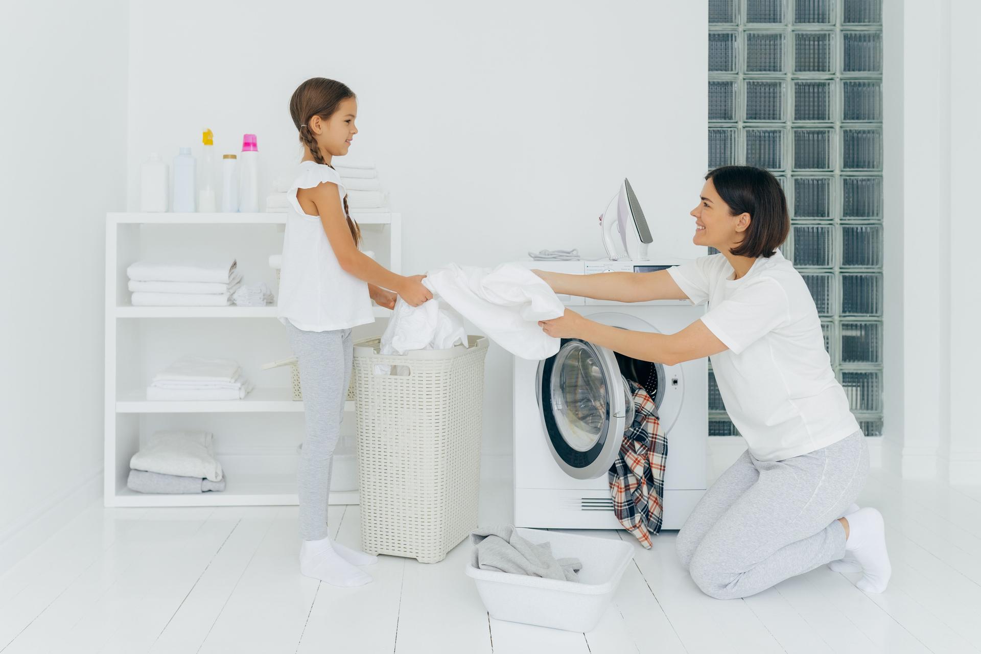 La mère heureuse charge des vêtements dans la machine à laver, la petite fille aide, donne le linge blanc du panier, font le ménage ensemble, rectifié dans des vêtements occasionnels. La fille offre l'aide à la maman, vêtements propres